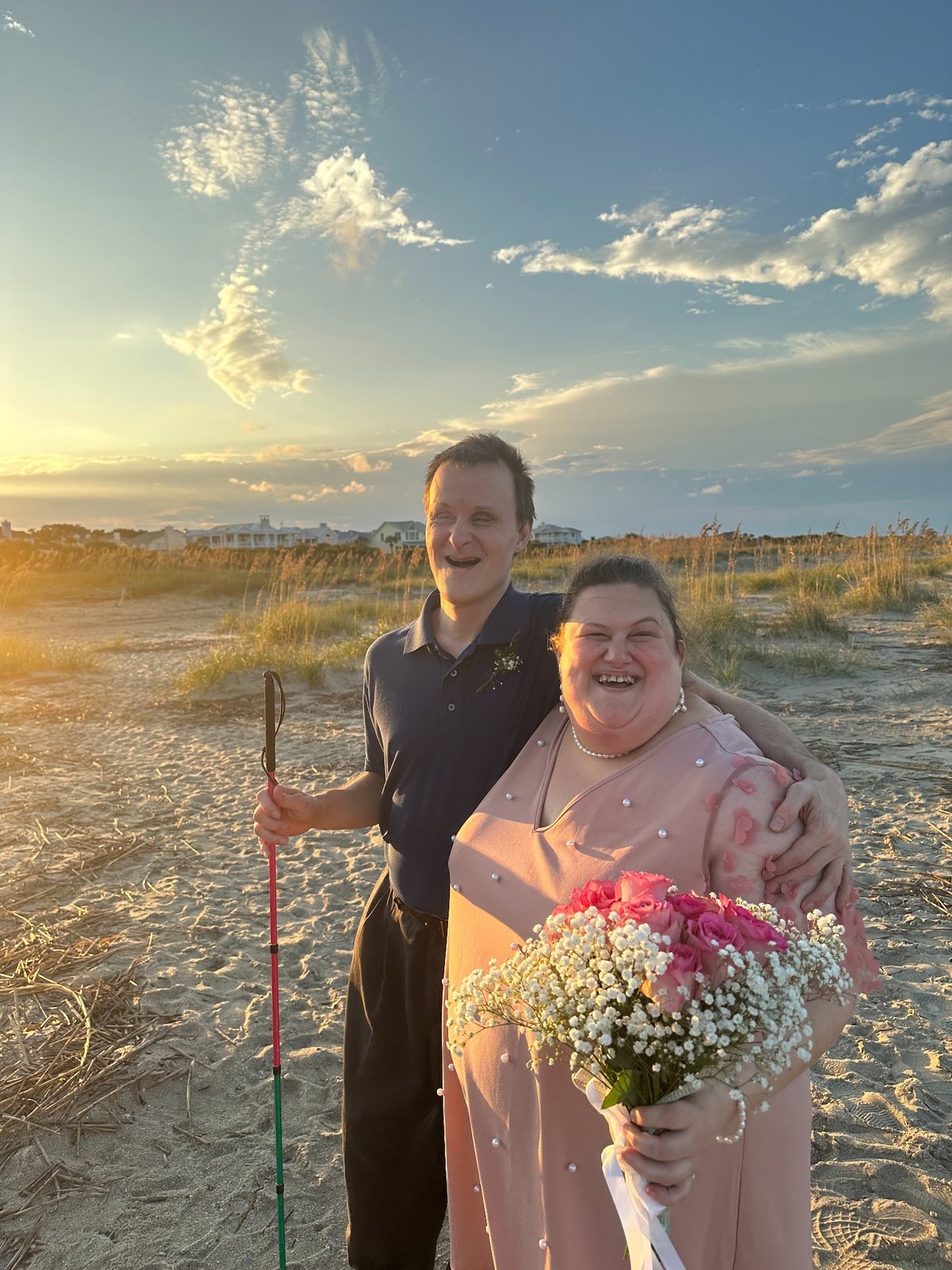 Jody and Amy smiling together on a beach at sunset on their wedding day, with Amy holding a bouquet of pink roses and baby’s breath.